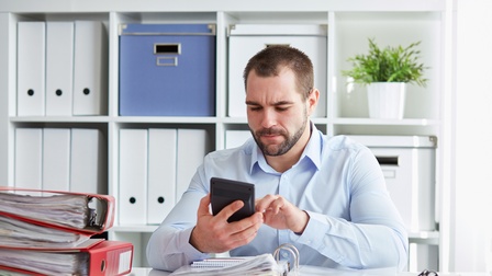 Frontale Aufnahme einer Person in einem hellblauen Hemd, die an einem Schreibtisch sitzt und auf einem Taschenrechner tippt. Vor ihr liegt ein aufgeklappter Ordner mit Unterlagen. Daneben sind weitere, geschlossene, dicke Ordner