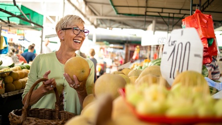 Seitliche Aufnahme einer Person, die in einem Supermarkt vor verschiedenen Obstsorten wie Bananen oder Mandarinen steht und in der linken Hand einen Granatapfel hält. Um die linke Schulter hat sie einen Einkaufskorb