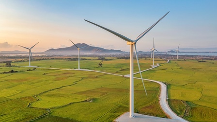 Landschaftsaufnahme einer grünen Landschaft, auf der versetzt Windräder stehen. Am Horizont ist eine Ortschaft vor einem Berg und neben einem Gewässer. Der Himmel ist blau und es herrscht Sonnenschein
