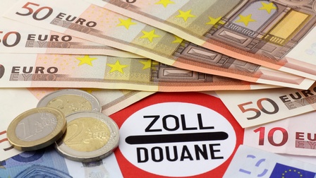 Nahaufnahme des Wortes Zoll in einem roten Kreis auf einem Tisch. Um den roten Kreis liegen verschiedene Euroscheine und Euromünzen