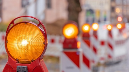 Aufnahme rot-weißer Absperrungen hintereinander auf der Straße. Auf jeder Absperrung ist oben ein oranges Licht montiert