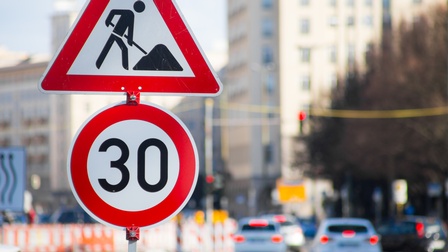 Zwei Straßenschilder im Fokus: Ein Baustellen- und ein 30-er-Zonene-Schild, im Hintergrund verschwommen Autos in Rückansicht.