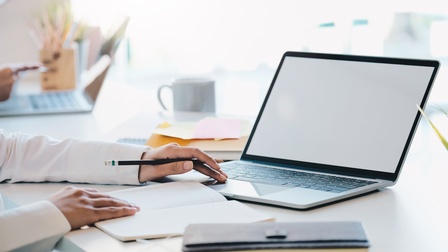 Eine Person sitzt an einem Tisch und hat vor sich ein aufgeklapptes Notizbuch. Daneben sind ein aufgeklappter Laptop, ein Buch sowie eine Tasse