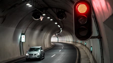 Eine rote Verkehrsampel ist an der Wand eines Straßentunnels angebracht. Die Fahrbahn verläuft in einer Kurve durch den Tunnel. Auf der Straße fährt ein Auto. Entlang der Tunnelwand befinden sich Beleuchtung und technische Einrichtungen