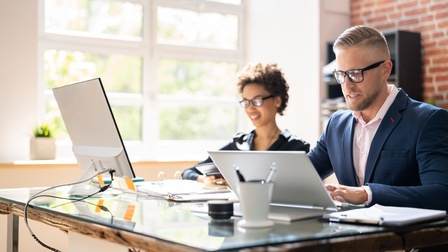 Zwei Personen mit Brillen sitzen an Schreibtisch vor Computermonitoren und blicken darauf, ringsum Büroutensilien, im Hintergrund verschwommen weißes Fenster und rote Ziegelwand