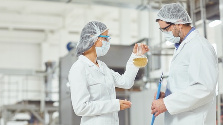 Zwei Personen in weißen Kitteln, mit Mundnasenschutz, Schutzbrillen und Haarnetzen stehen einander gegenüber; eine Person hält Glasgefäß mit gelblicher Flüssigkeit hoch, auf das beide Personen blicken, die andere Person hält Clipboard in Händen