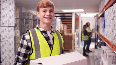 Lächelnde Person in gelber Schutzweste hält weißes Paket in Händen, im Hintergrund verschwommen weitere Personen in Gang einer Lagerhalle vor Regalen stehend