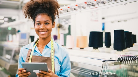 Lächelnde Person in Schürze mit Maßband um den Hals hält Tablet in Händen, im Hintergrund Stickmaschine mit aufgesteckten Garnen