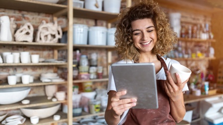 Person mit lockigem Haar und roter Arbeitsschürze hält eine Tasse und blickt freudig auf ein Tablet, im Hintergrund zeigt sich ein Regal mit Tonwaren und Arbeitsmaterialien