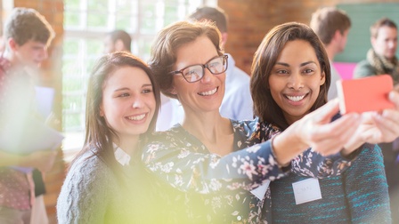 Person mittleren Alters mit Brille hält ein Smartphone und blickt gemeinsam mit zwei jugendlichen Personen darauf, im Hintergrund stehen weitere Personen in einem Raum
