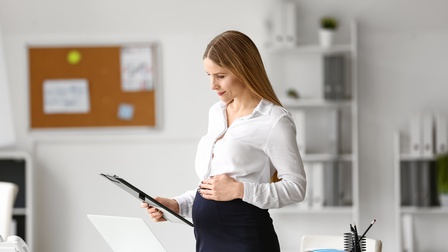 Eine Person in weißer Bluse und dunkelblauem Rock steht leicht seitlich zur Kamera. Sie blickt auf ein Clipboard, das sie mit der rechten Hand hält. Ihre linke Hand liegt auf ihrem gewölbtem Bauch. Direkt hinter ihr ist ein Schreibtisch mit Laptop