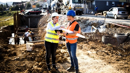 Zwei Personen in Schutzwesten und mit Helmen stehen bei einer Baustelle neben einem Erdloch. Im Erdloch sind mehrere Personen mit Helmen, die Holzbalken anbringen. Neben der Baustelle sind Wohnhäuser und Straßen