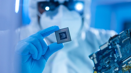 Person mit Anzug, Schutzmaske und Brille sowie blauen Schutzhandschuhen hält einen Prozessor in Händen