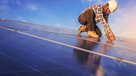 Person mit weißem Schutzhelm steht auf einem Dach. Auf dem Dach sind mehrere Solarpaneele, die jeweils einen Metallrahmen haben. Die Person hat ihre Hände auf einem Metallrahmen. Der Himmel ist blau
