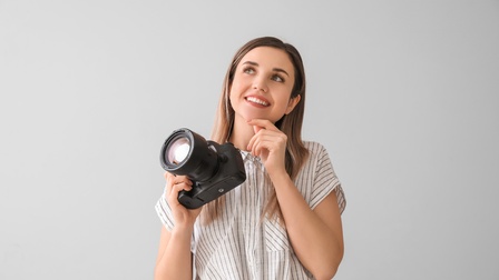 Eine Person in Frontalansicht steht vor einer hellen Wand und hält einen Fotoapparat in der rechten Hand. Die Person blickt nachdenklich nach oben