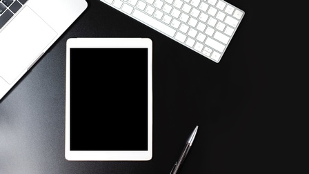 Mock-up von einem Tablett auf einem schwarzen Untergrund neben einem Laptop, einer Tastatur sowie einem Kugelschreiber, Topshot