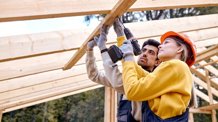 Zwei Personen stehen unter Holzbalken, die quer durch das Bild verlaufen. Die linke Person hält da Brett mit beiden Händen. Die rechte Person mit einer Hand. In der linken hält sie eine Bohrmaschine gegen das Brett. Hinter ihnen sind Bäume.