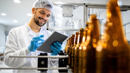 Auf einem Förderband stehen hintereinander in einer Reihe viele dunkle Glasflaschen. Links daneben steht eine Person mit Haarnetz, Handschuhen und Tablet, die auf das Tablet blickt