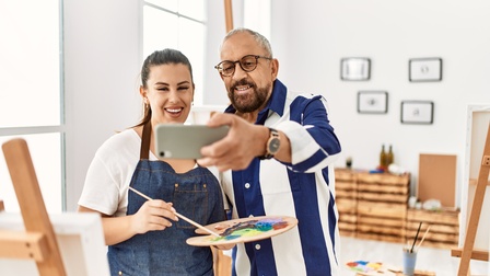 Zwei Personen stehen nebeneinander in einem hellen Raum. Die rechte Person macht mit einem Handy ein Selfie von ihnen. Die linke Person hält einen Pinsel. Vor ihnen steht eine Staffelei mit einer Leinwand