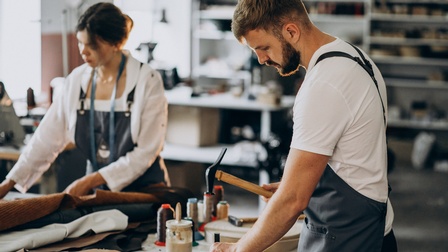 Eine Person in einer Schürze steht an einem Tisch in einer Werkstatt und bearbeitet mit einem Hammer ein Stück Leder. Auf dem Tisch sind verschiedene Lederstoffe. Am Tisch steht eine weitere Person mit Schürze