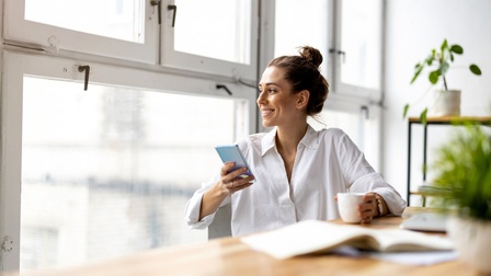 Lächelnde Person blickt aus Fenster an Tisch sitzend mit Smartphone in der Hand