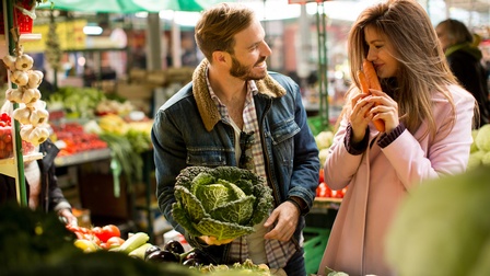 Zwei Personen stehen an Gemüsestand auf Markt, eine Person hält Kohlkopf in Händen, die andere riecht an einem Bund Karotten