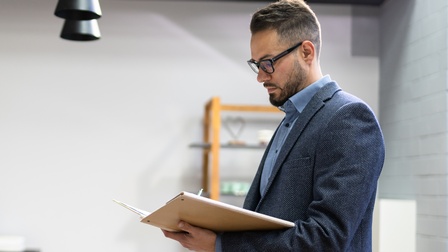 Person mit dunklen kurzen Haaren, Bart, Brille, Anzug steht und blickt auf eine Dokumentenmappe