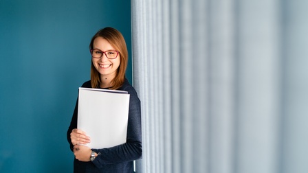 Person mit langen braunen Haaren und Brille hält Unterlagen und steht freudig vor einer blauen Wand und einer geschlossenen Fensterfront mit grauen Lamellenvorhang