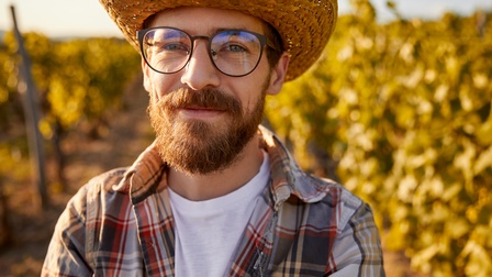 Bärtige Person mit Brillen und Strohhut im Porträt, im Hintergrund verschwommen grüngold glänzende Weinreben