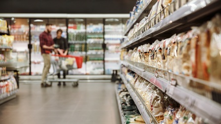 Rechts im Fokus ist ein Regal im Gang eines Supermarkts, das mit verschiedenen Produkten befüllt ist. Im Hintergrund stehen zwei Personen neben einem teilweise gefüllten Einkaufswagen. Hinter ihnen ist ein Kühlregal