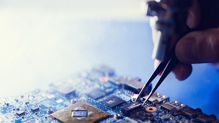 Nahaufnahme einer elektronischen Leiterplatte mit verschiedenen Mikrochips und Anschlüssen. Eine Hand hält eine Pinzette über einem kleinen elektronischen Bauteil. Mehrere Komponenten und Leiterbahnen sind auf der Leiterplatte