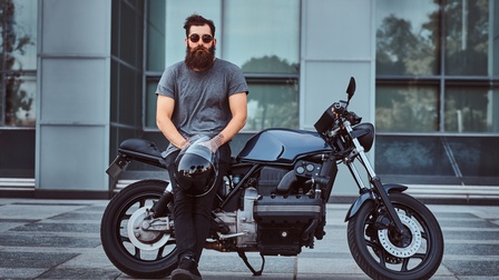 Eine Person lehnt seitlich an einem stehenden, schwarzen Motorrad. Die Person hält einen schwarzen Helm in den Händen. Das Motorrad steht auf einem Boden mit großen Fliesen. Im Hintergrund ist ein Gebäude.