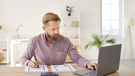 Frontale Aufnahme einer Person, die an einem Tisch sitzt und auf einen aufgeklappten Laptop blickt. Vor der Person sind nebeneinander zwei Stapel Papiere. Daneben steht ein Taschenrechner