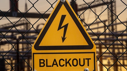 Nahaufnahme eines gelben Schildes in Dreiecksform an einem Maschendrahtzaun befestigt, der Rand in Schwarz in der Mitte Symbol eines nach unten deutenden Blitzes. Darunter weiteres Schild mit Schriftzug Blackout.