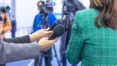 Rückenansicht einer Person in einem grünen Sakko und mit offenen, braunen Haaren. Links neben der Person halten drei Personen jeweils ein Mikrofon. Im Hintergrund stehen zwei Personen hinter Filmkameras