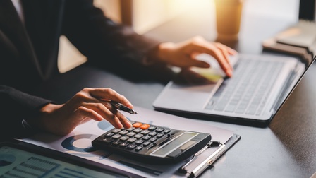 Hände einer Person in Nahaufnahme. Mit der linken Hand tippt sie auf einem Laptop, mit der rechten Hand auf einem Taschenrechner.