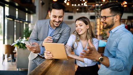 Drei lächelnde Personen stehen in Lokal und blicken auf Tablet, das eine Person hält, eine Person hält Smartphone in einer Hand