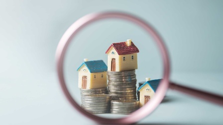 Blick durch ein Lupenglas auf Stapel von Münzen, auf denen kleine Häuser stehen