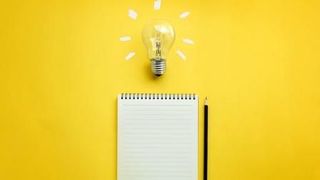 Auf gelbem Untergrund liegt ein Notizblock. Rechts neben dem Notizblock ist ein Bleistift. Über dem Notizblock ist eine Glühbirne mit weißen Strichen, die von ihr weggehen