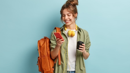 Jugendliche Person mit braunen hochgeschlossenen Haaren, weißem Shirt und grüner offener Bluse blickt auf ihr Smartphone und trägt einen orangen Rucksack sowie gelbe Kopfhörer rund um den Hals und einen Kaffeebecher in der Hand