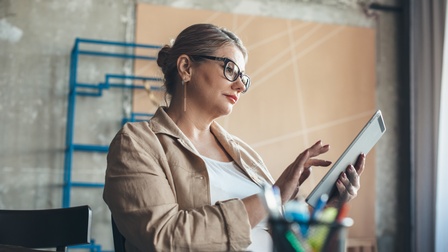 Person mit geschlossenen grau-braunen Haaren, Brille und Ohrringen sowie weißem Shirt und beiger Jacke hält ein Tablet in den Händen und blickt darauf, im Hintergrund zeigt sich ein ein minimalistischer Raum in Betonoptik mit beiger Leinwand