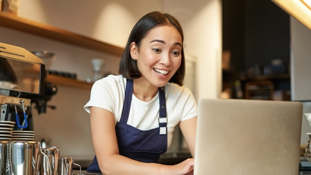 Person in Arbeitskleidung mit blauer Schürze steht in einem Café an der Theke und blickt freudig in einen Laptop