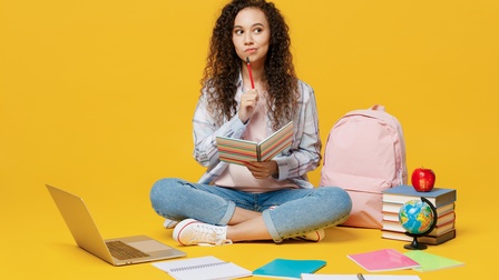 Person sitzt in Schneidersitz am Boden, in einer Hand Notizbuch haltend, andere Hand mit Stift an Kinn haltend, ringsum Notizbücher, Bücherstapel, Globus, Apfel, Rucksack und aufgeklapptes Notebook, Hintergrund gelb