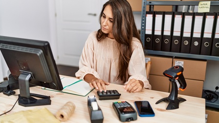Person sitzt an einem Schreibtisch und blickt auf ihren Bildschirm, während sie einen Taschenrechner bedient, im Hintergrund steht ein Regal mit Ordnern und Kartonagen sowie stehen am Schreibtisch noch ein Scan- und ein digitales Kartenbezahlgerät, ein Smartphone und Verpackungsmaterialien