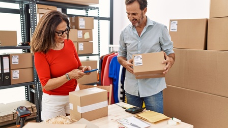 Zwei Personen stehen an einem Tisch. Die rechte Person hält in den Händen ein kleines Päckchen. Die linke Person hält ein Smartphone, das sie über ein am Tisch stehendes Paket hält. Im Hintergrund stehen weitere Kartons