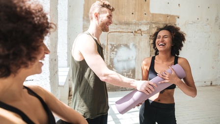 Drei lächelnde Personen in Gymnastikkleidung stehen in Halle mit kaum verputzten Wänden, eine Person hält zusammengerollte lavendelfarbene Gymnastikmatte andere Person mit Bart greift danach