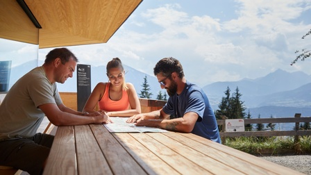 Drei Personen sitzen an Holztisch im Freien und blicken auf Karte, im Hintergrund Holzzaun, Wiese, Bäume und Gebirgszug