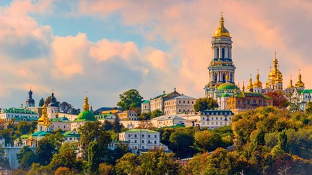 An einem Hügel liegen viele verschiedene Gebäude sowie Kirchen. Die Kirchen haben teilweise mehrere Türme. Die Dächer der Türme sind golden. Daneben sind historische Gebäude. Vor den Gebäuden sind grüne Bäume