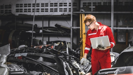 Person in rotem Arbeitsoverall mit Schutzbrillen und Handschuhen hält aufgeklapptes Notebook in einer Hand und sichtete umherliegende Autoteile in Halle