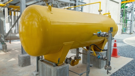 Ein großer, gelber Tank steht im Freien. Dahinter ist eine Industrieanlage mit Metallstangen